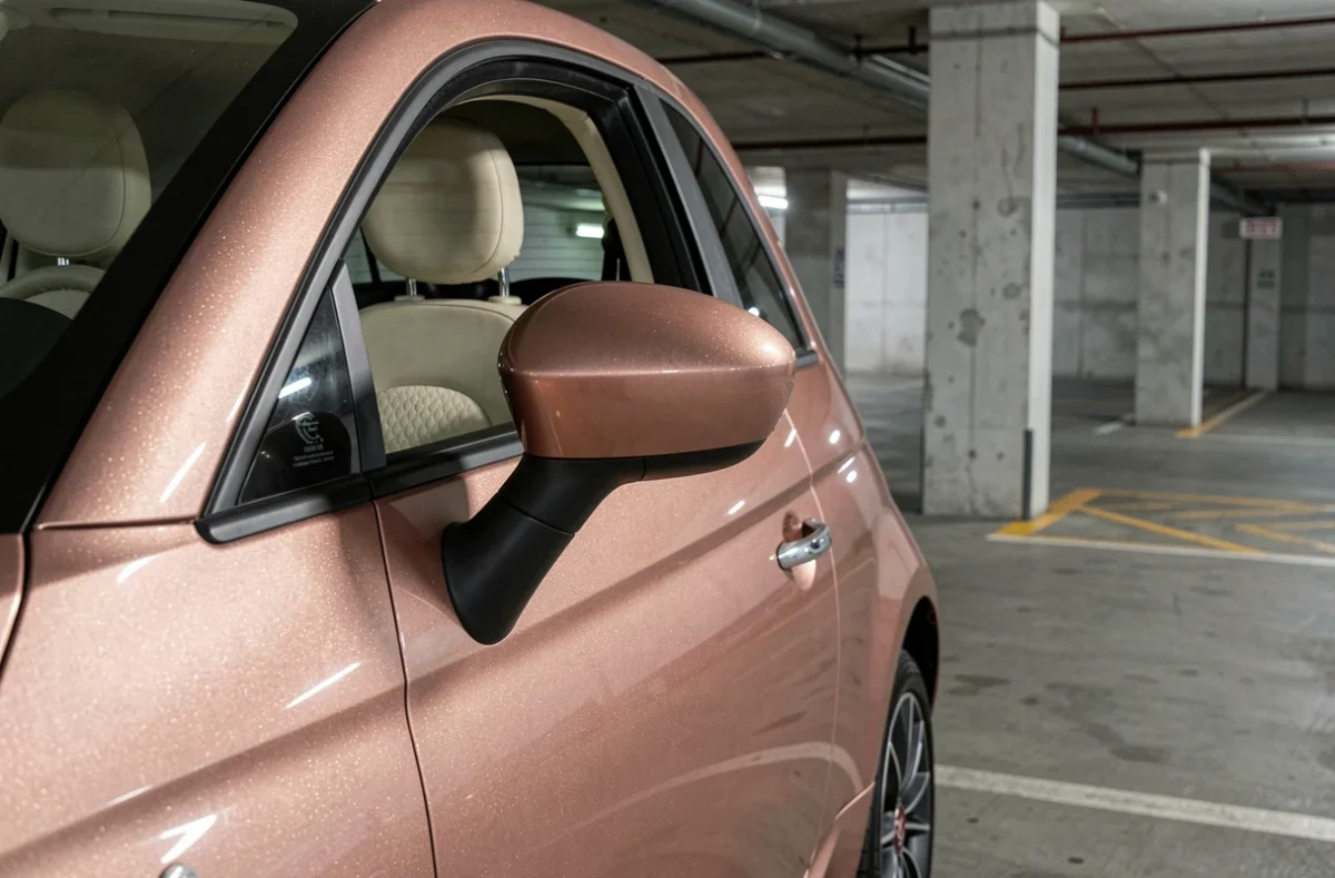 Détail de la poignée et du rétroviseur d'une Fiat 500 rose gold avec intérieur cuir crème visible, dans un parking souterrain